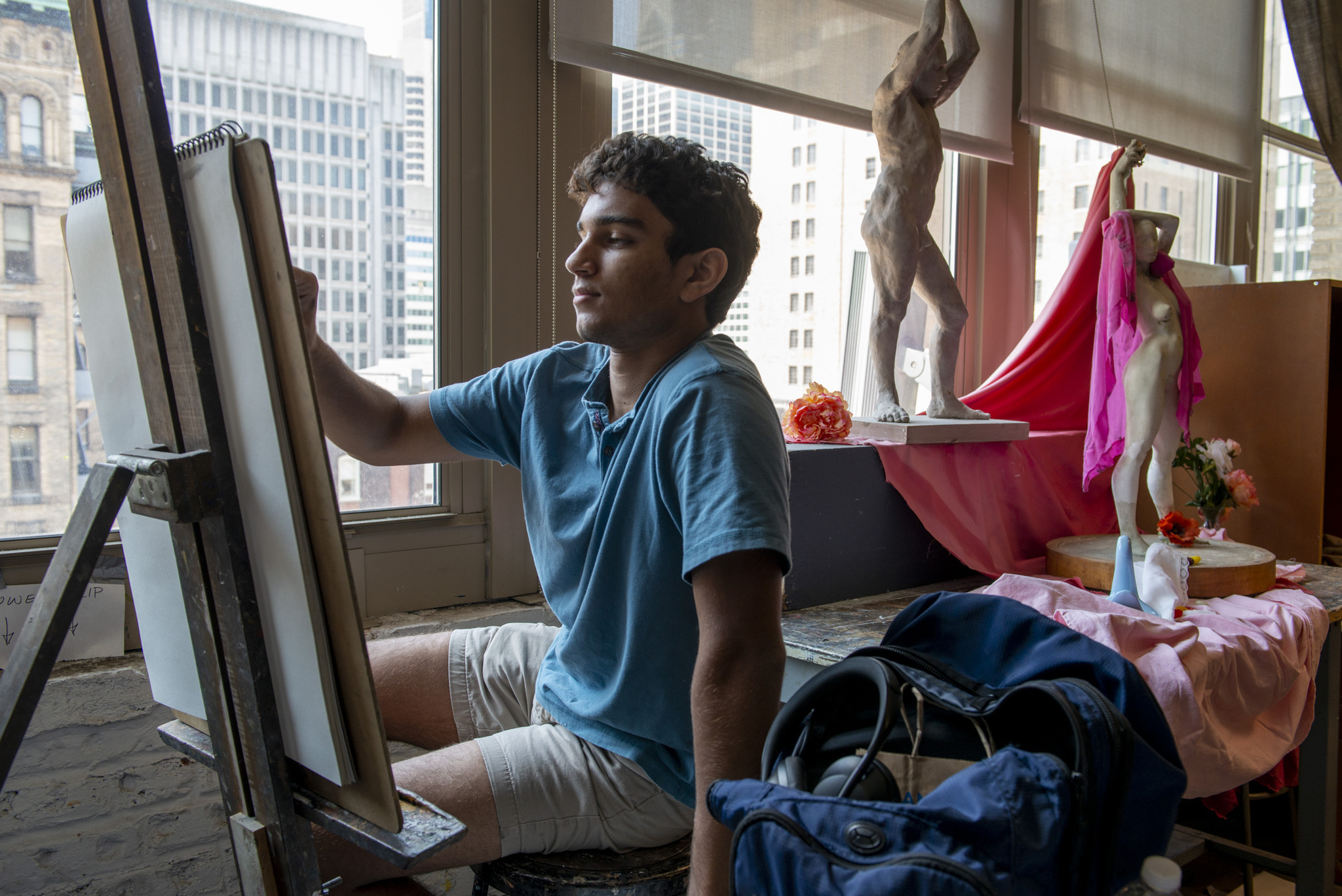 A student in a blue tee shirt reaches up to draw on a pad of paper that leans on an easel. The easel is turned away from the camera. In the background, a table is draped with pink fabric, plaster casts of figures, and other still life objects. Windows behind the table reveal tall buildings outside.