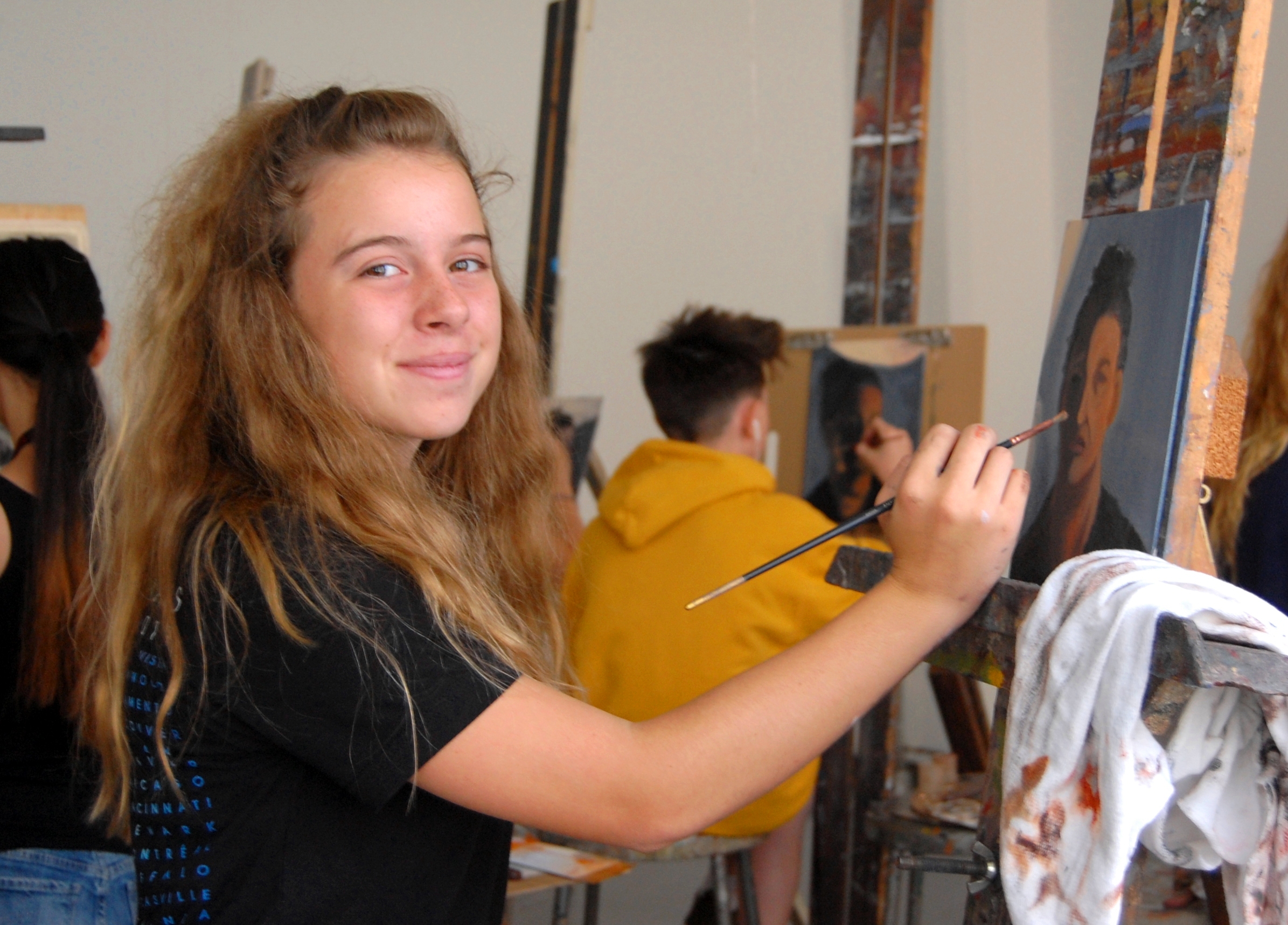 A student with long blonde hair smiles at the camera as she holds a paintbrush up to her easel, on which there is a portrait. In the background, another student in a yellow hoodie is painting a portrait.