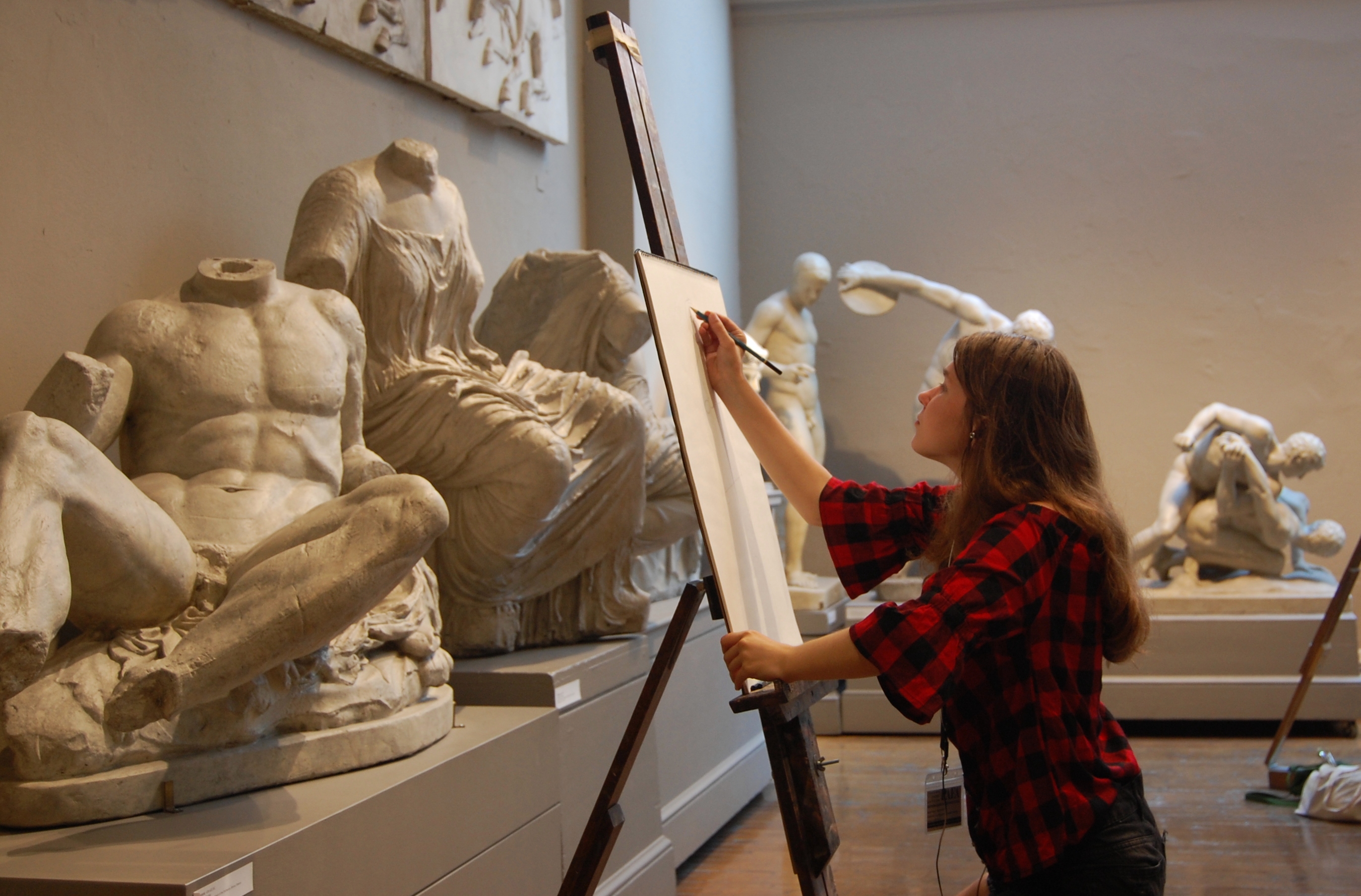 A student in a red and black plaid shirt draws on an easel in a room full of white plaster casts of classical statues