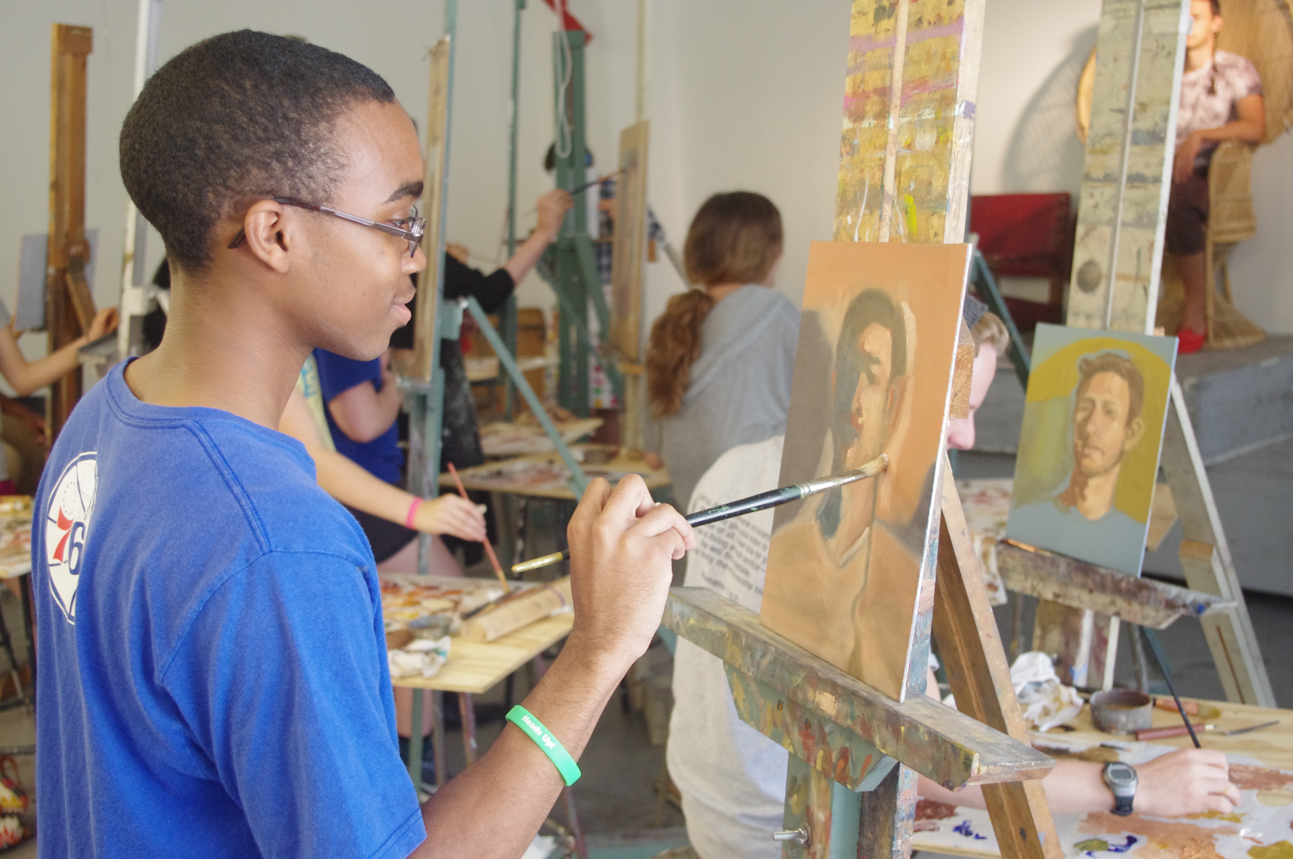 A student in a blue shirt holds a paintbrush up to an in-progress portrait on an easel. Other students work on easels in the background.