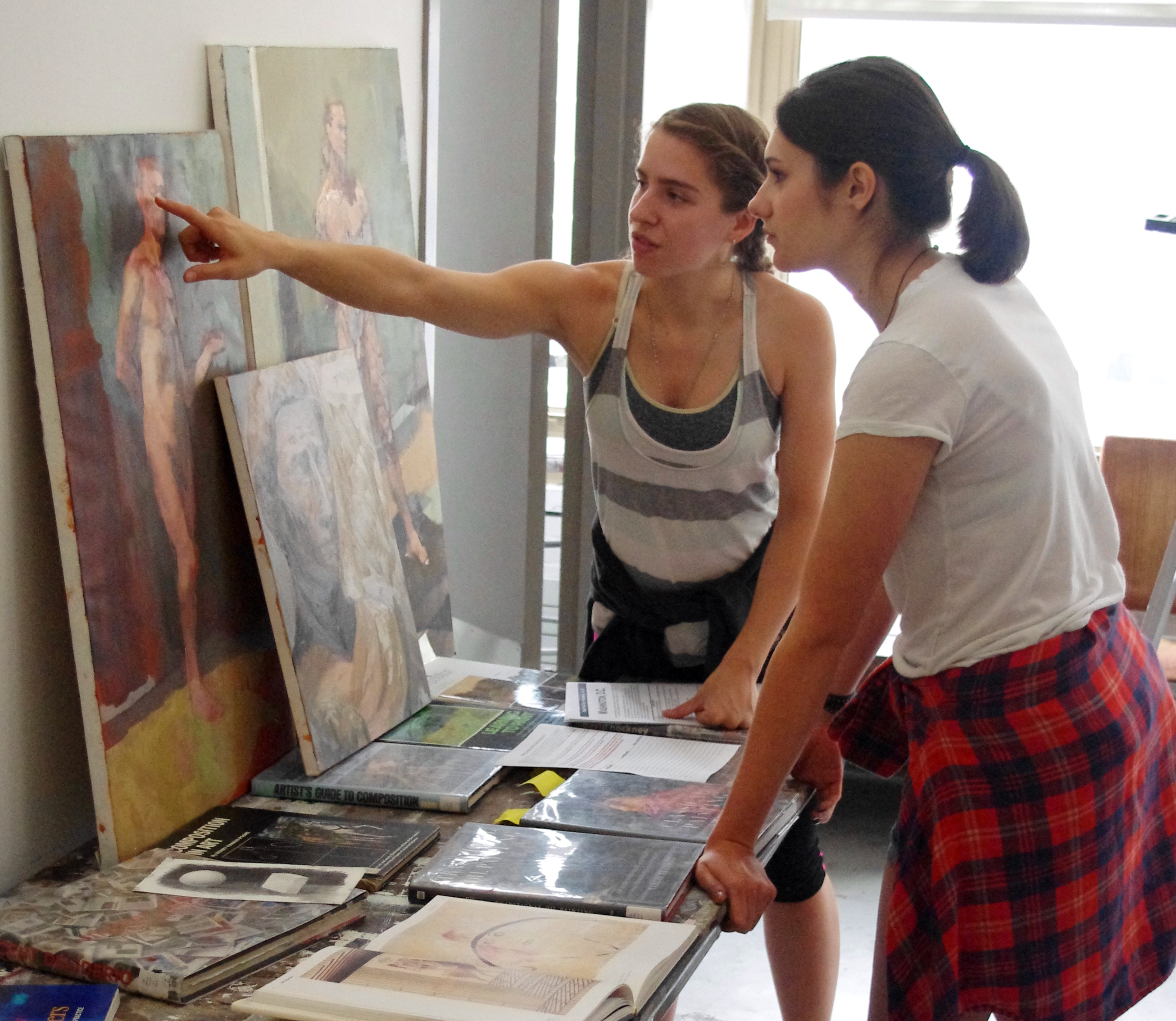 An instructor and a student look at a collection of artwork on a table. Three paintings lean against the wall, and the instructor points to one of the paintings.