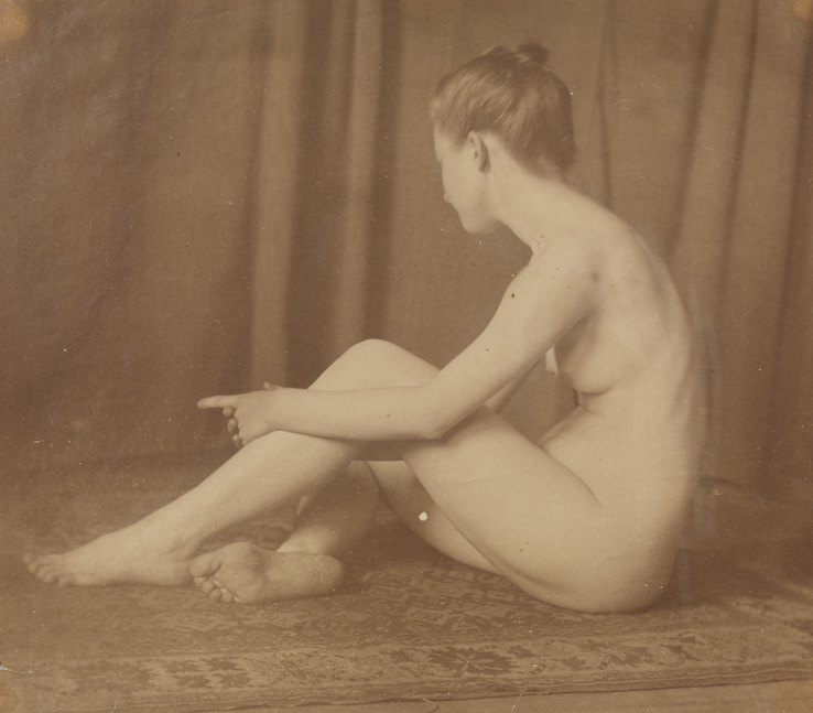 Female nude sitting on floor, grasping left leg, index finger pointed