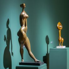 installation photo of two bronze works by John Rhoden on pedestals. The gallery and pedestals are painted green. The work in the foreground, "Eve", is a nude female figure in a dance position holding a ball in the right hand. The piece in the background is, Keesong, an abstract figure. . 