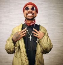 Portrait of Dave Hem smiling, wearing round sunglasses, a red knit skull cap and bandana, a yellow jacket and black shirt, with a big cross on a chain. Hem is showing the back of his hands, with three fingers on each extended toward each other. 