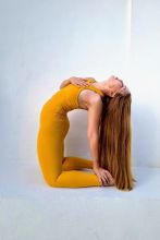 Photo of Eunmi Chang in a yoga pose starting on the knees with body arching back and hands connecting to the feet. Eunmi is wearing a mustard yellow leotard and has very long straight reddish-blond hair. 