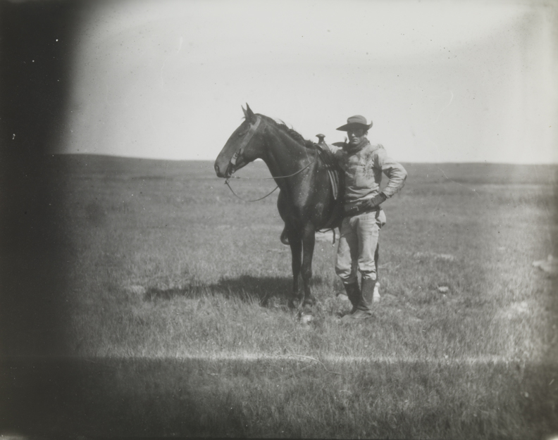 Cowboy in buckskin shirt, standing next to dark horse on grassland