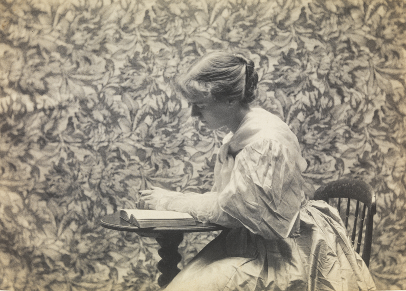 Eva Watson reading at small round table in front of floral backdrop