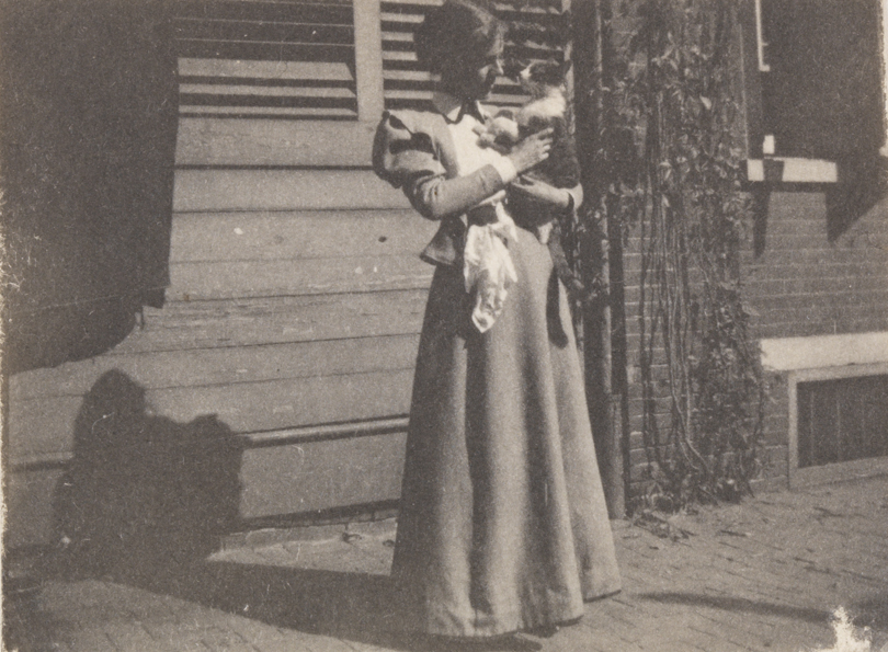 Eva Watson (?) holding grey tiger-striped cat in front of house