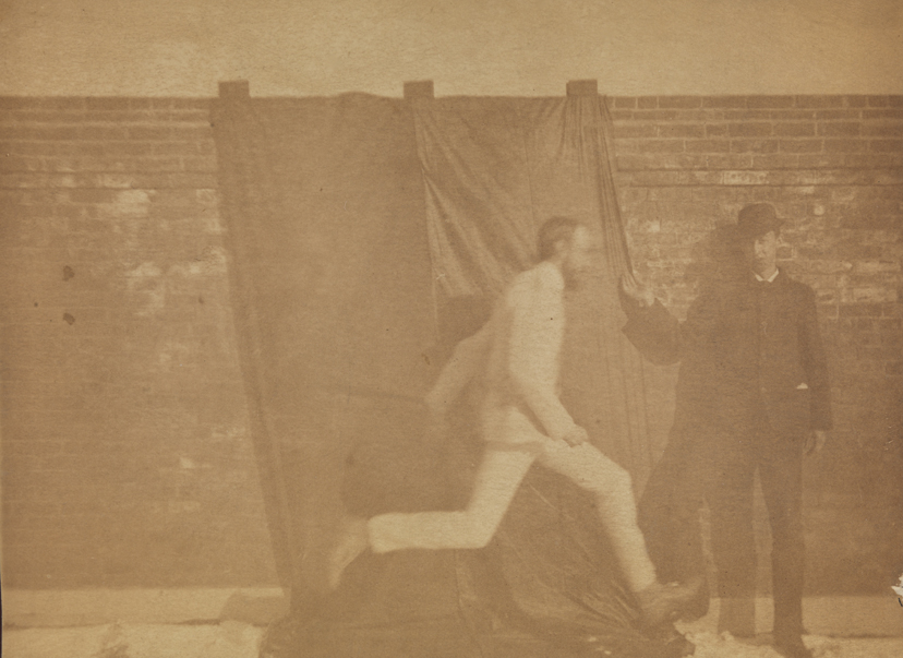 Motion study: man in light suit running in front of draped brick wall; man in dark suit standing at right