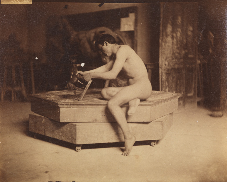 Male nude sitting on modeling stand, holding small sculpture of horse, in Pennsylvania Academy studio