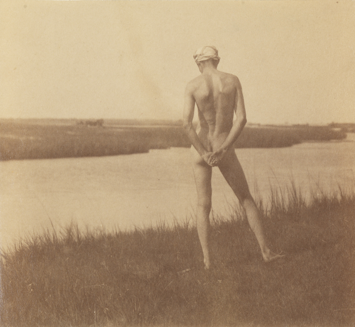 Male nude wearing head scarf, from rear, at edge of river