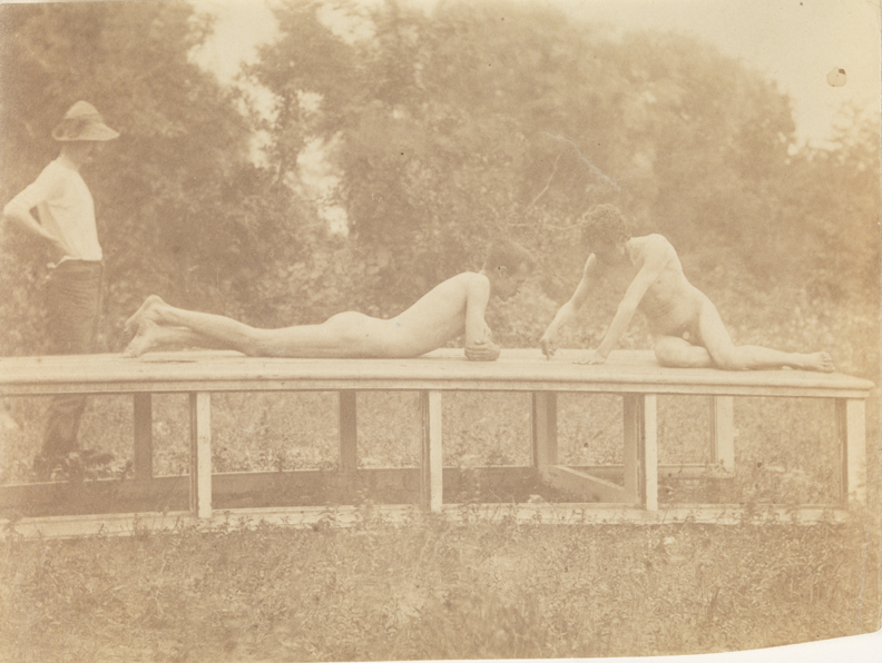 Male clothed, standing, and male nude and J. Laurie Wallace nude, reclining on platform in wooded landscape