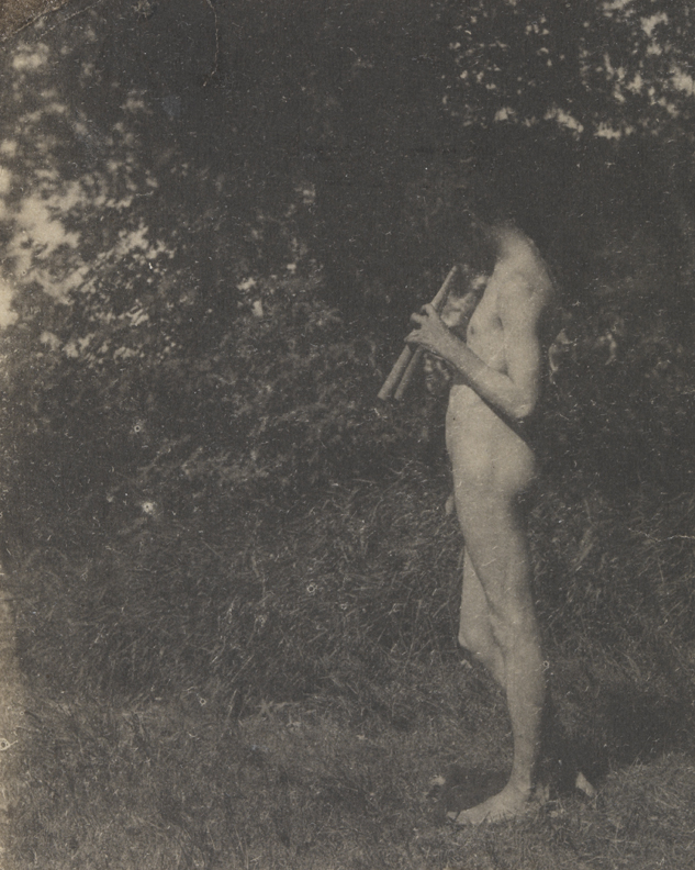 J. Laurie Wallace nude, playing pipes, facing left, in front of leafy background