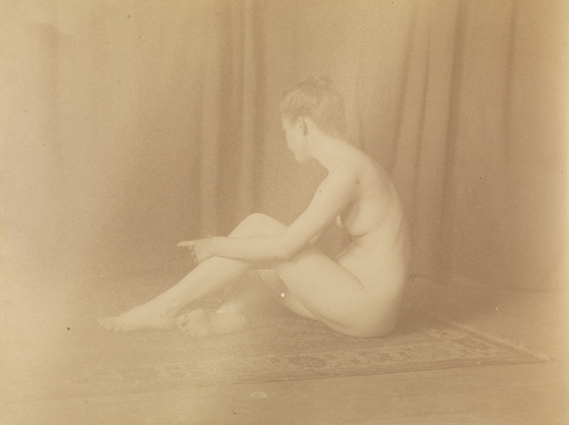 Female nude sitting on floor, grasping left leg, index finger pointed