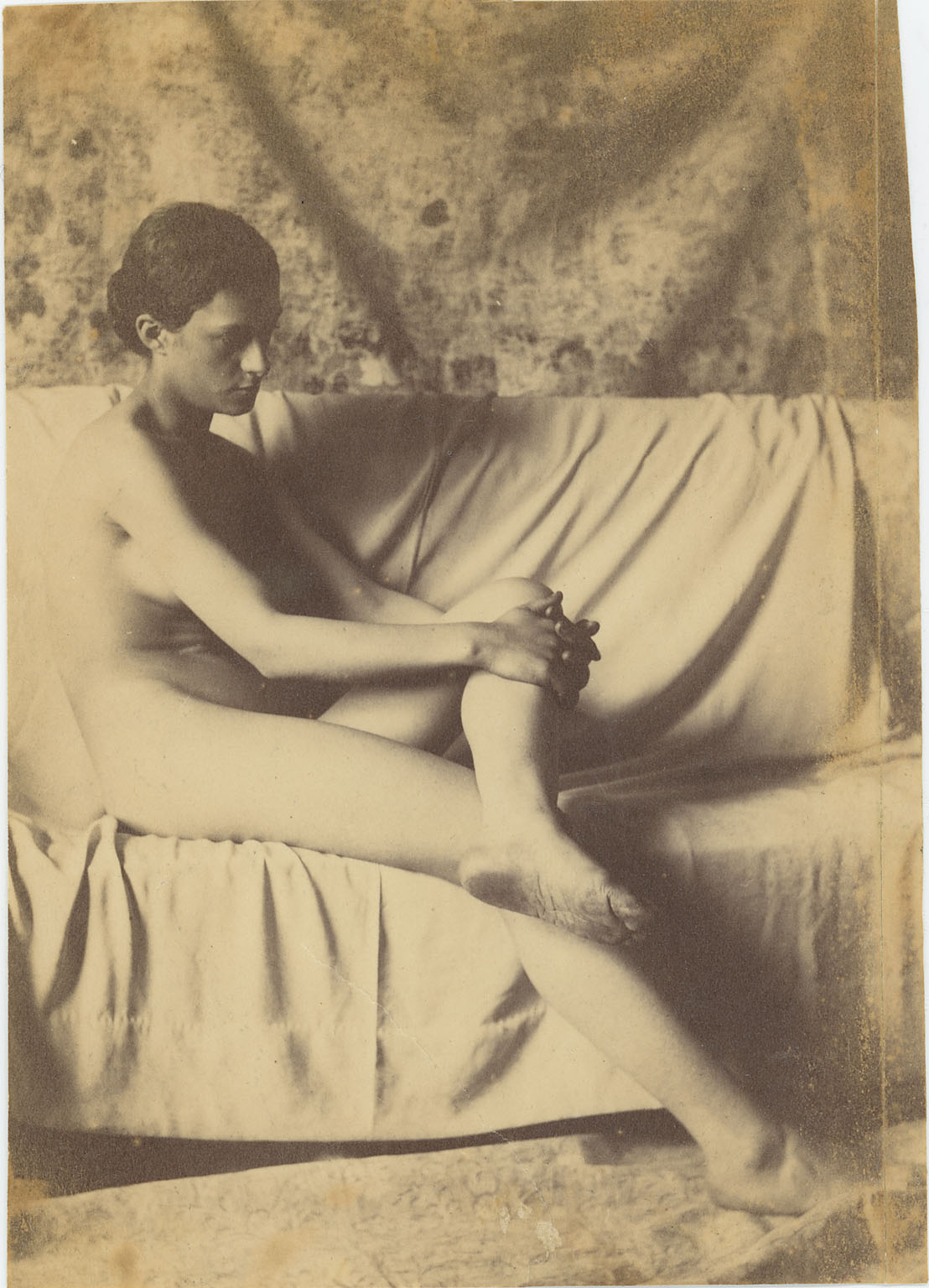 Female nude, sitting on draped couch, hands clasped around knee