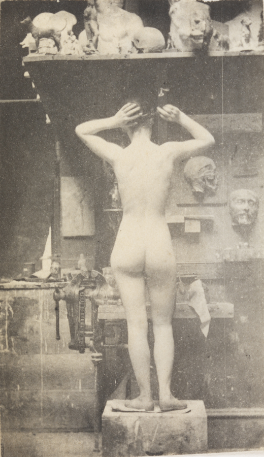 Female nude standing on wooden block, arms raised, from rear