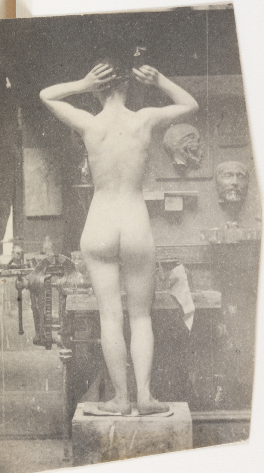 Female nude standing on wooden block, arms raised, from rear