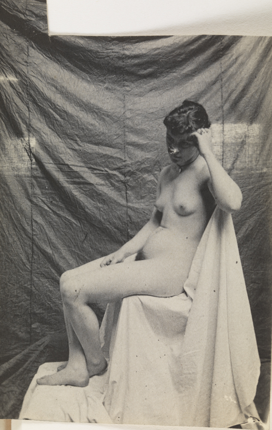 Female nude sitting on cloth-draped chair, facing left
