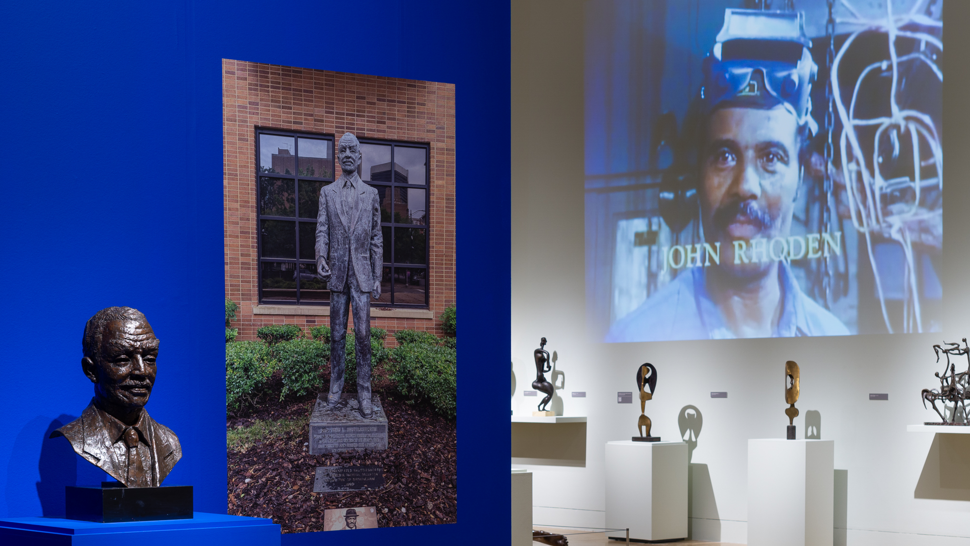 Installation view of the bust of Fred Shuttlesworth with an image of the full sculpture in the background, and to the right are more sculptures on view.