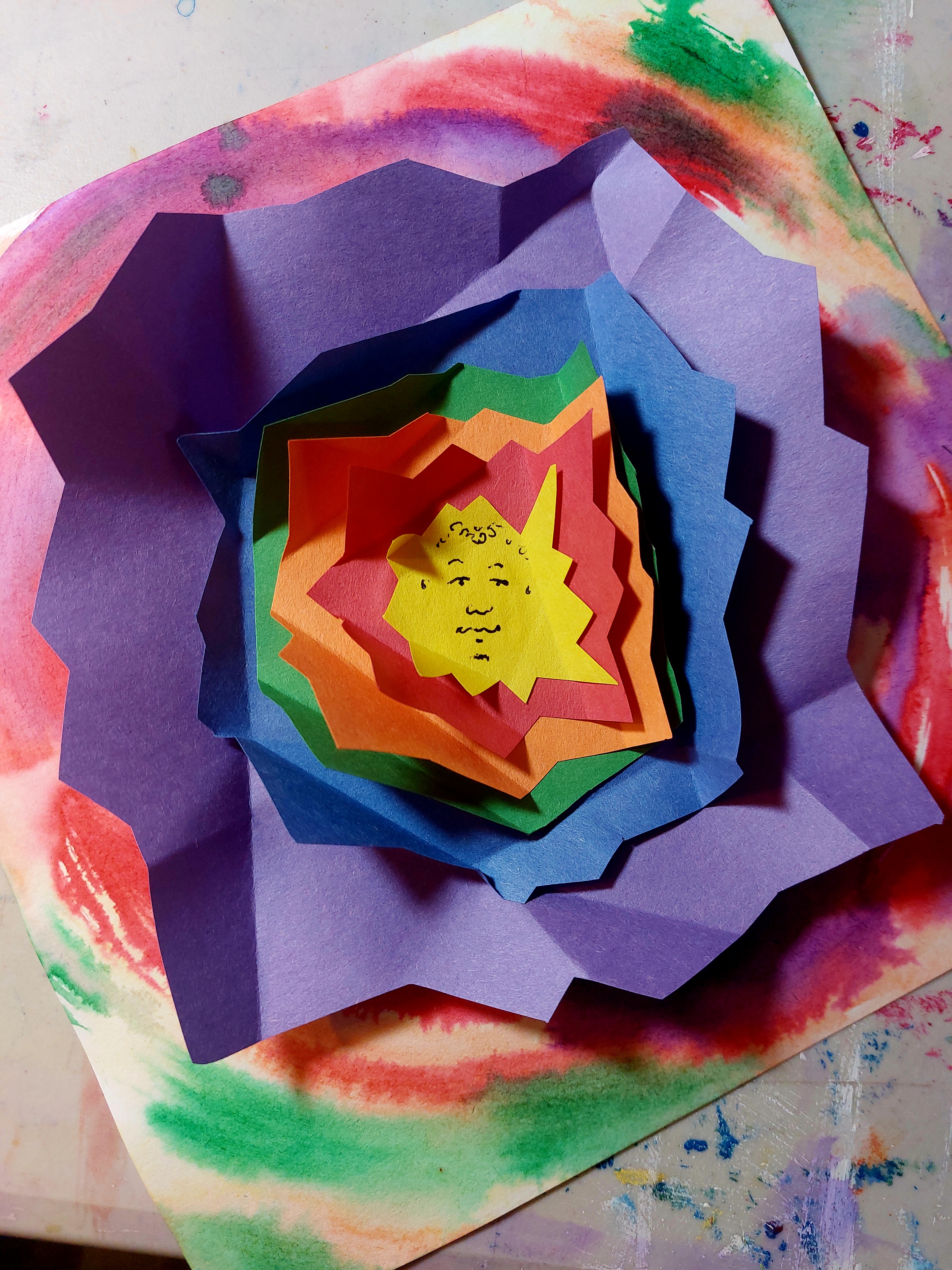 Layered, colorful paper art forms a star shape with a small, simple face in the center. The background is a vibrant swirl of red, green, and purple.