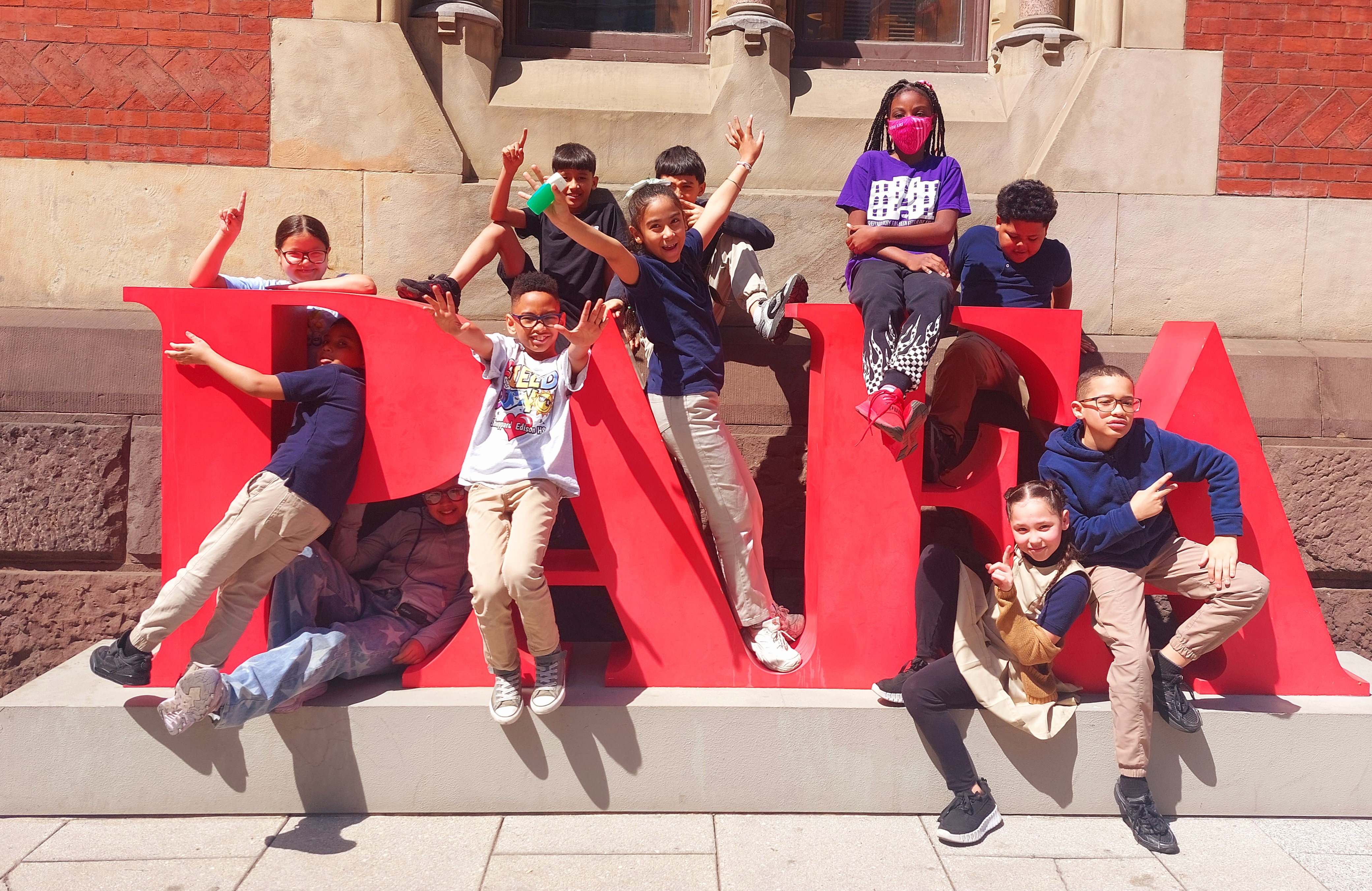 A group of joyful children poses playfully around large red "PARA" letters. They wear casual clothes and smile brightly, exuding excitement and camaraderie.