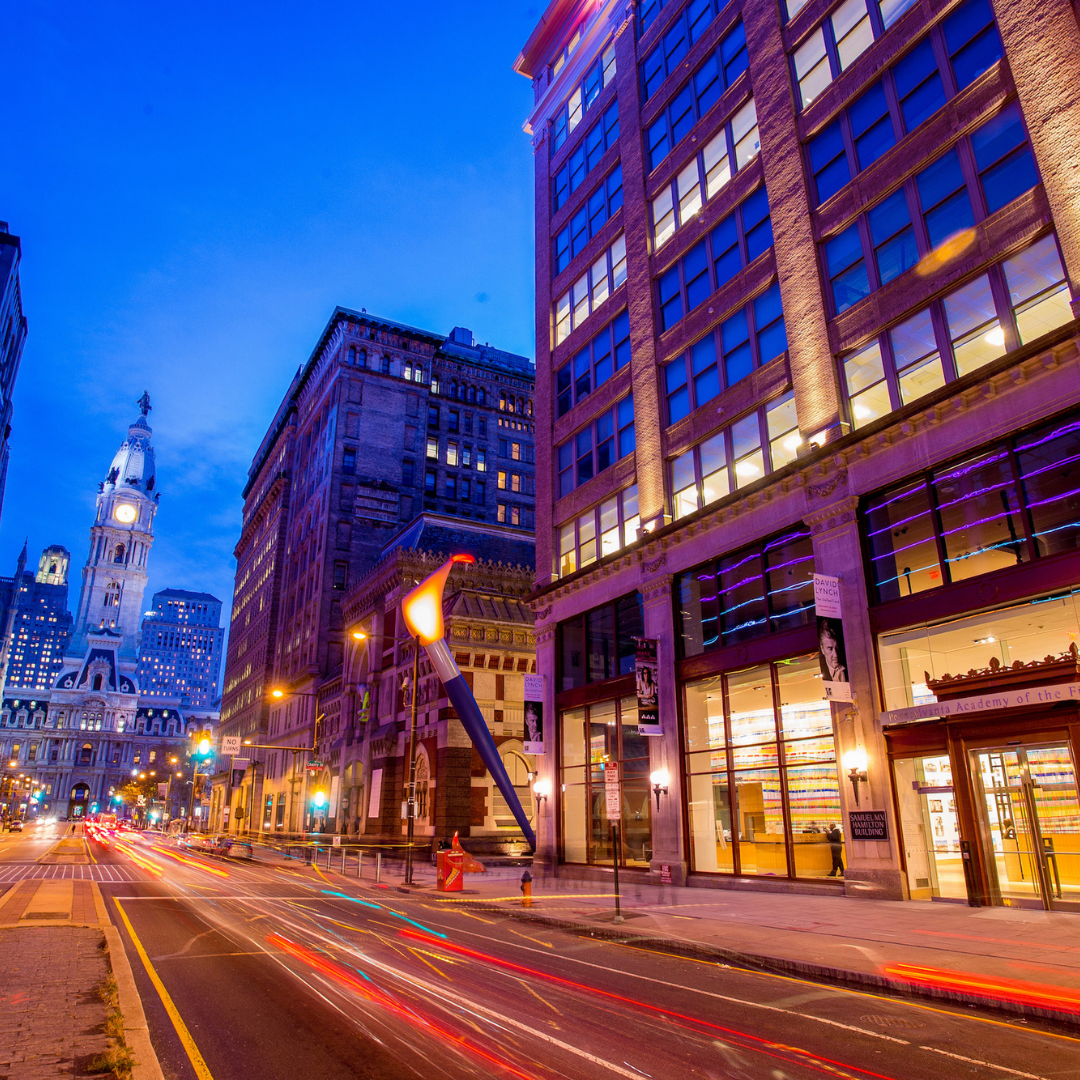 A nighttime street view features a large paintbrush sculpture standing outside a multi-story building, with light trails from cars illuminating the road and a historic clock tower visible in the distance.