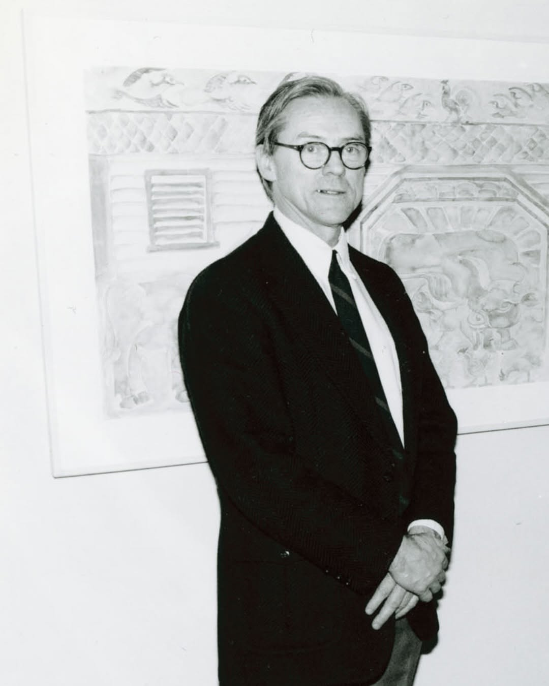 A black and white portrait of a man with short, combed hair and glasses, wearing a dark suit jacket, a white collared shirt, and a dark necktie, standing with his hands clasped in front of a framed piece of artwork.