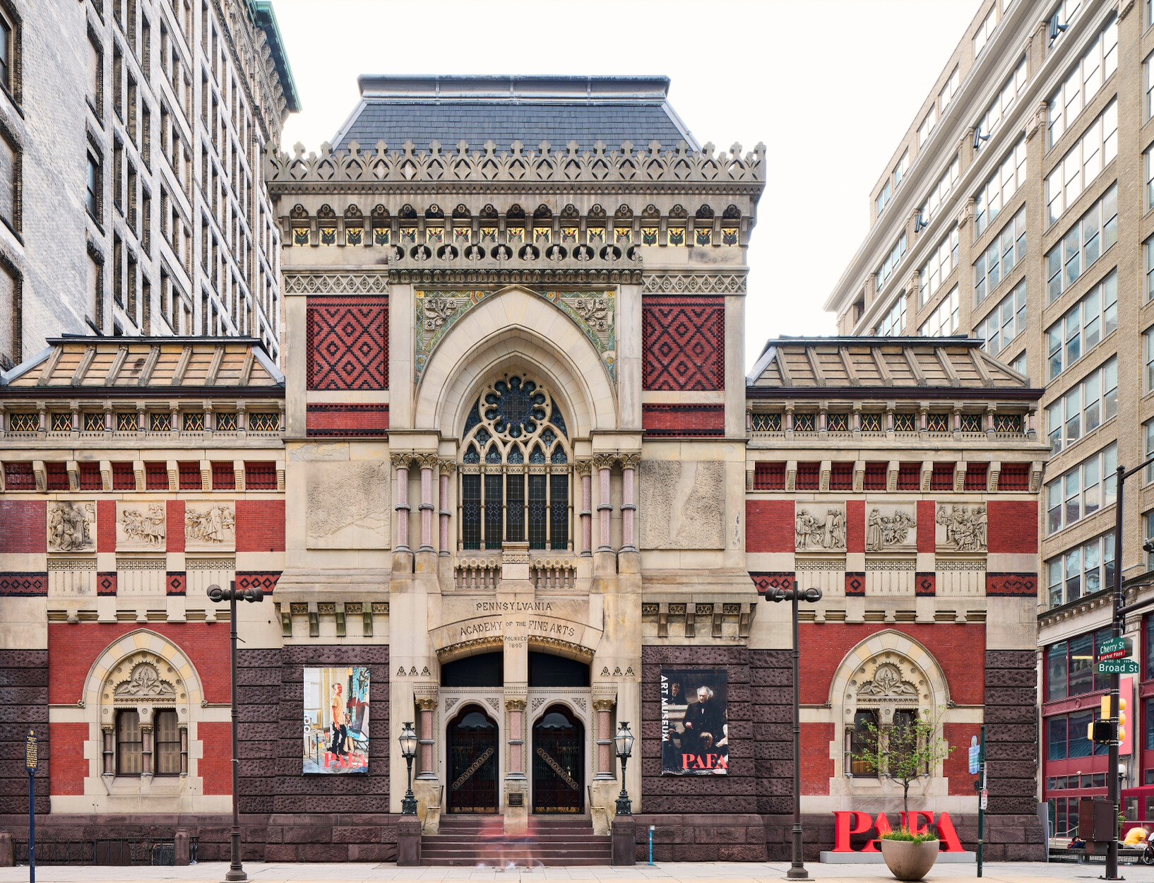 Ornate, historic building facade with Gothic arches, detailed brickwork, and colorful patterns. Flanked by modern urban architecture, conveying a blend of old and new.