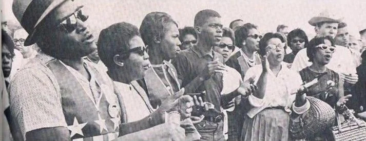 A black and white photo of a group of people singing passionately at a civil rights protest. The crowd exudes unity and determination.