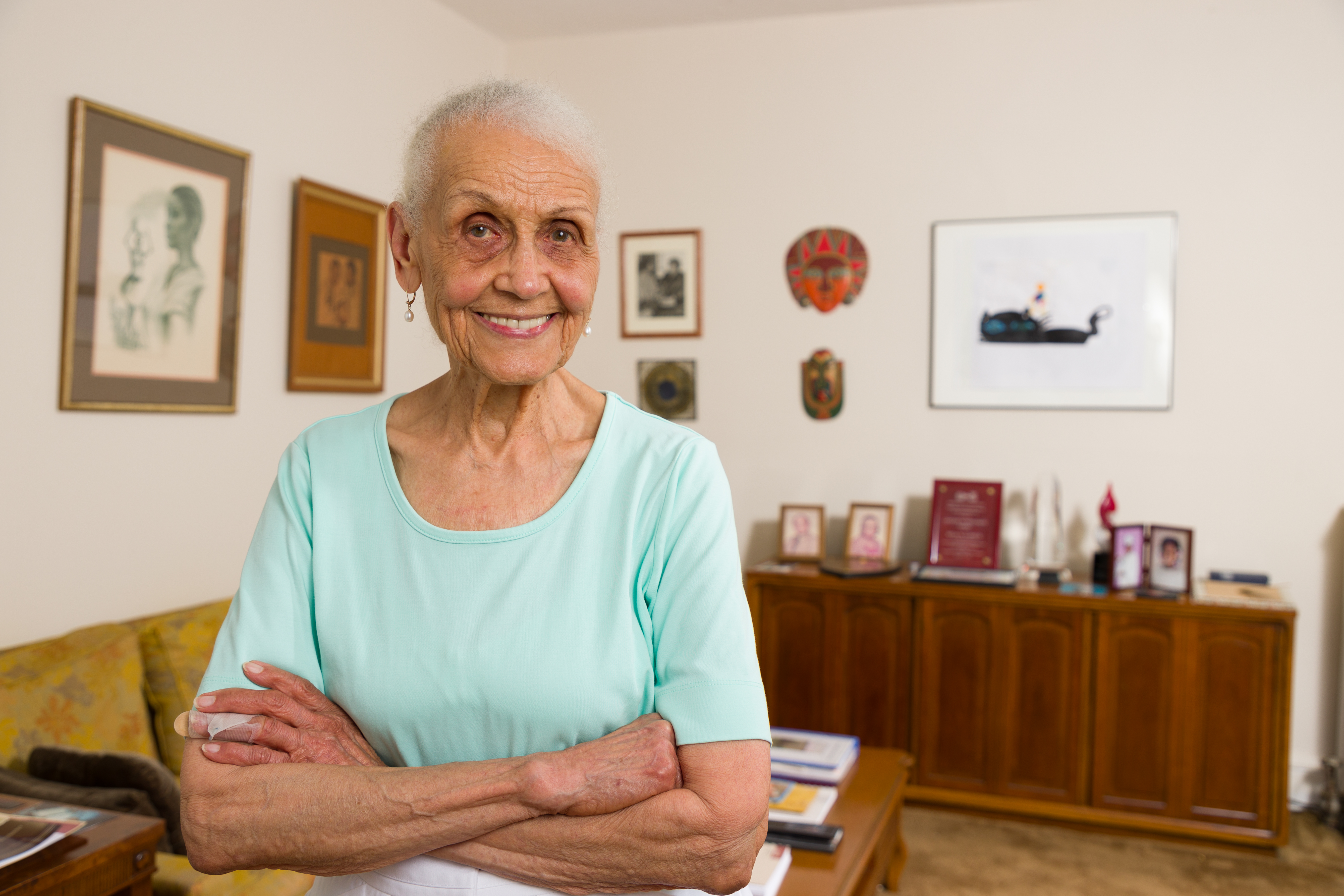 Elderly woman with short hair and a warm smile stands confidently in a cozy room with art on the walls and books on a wooden cabinet, conveying a welcoming and homely atmosphere.