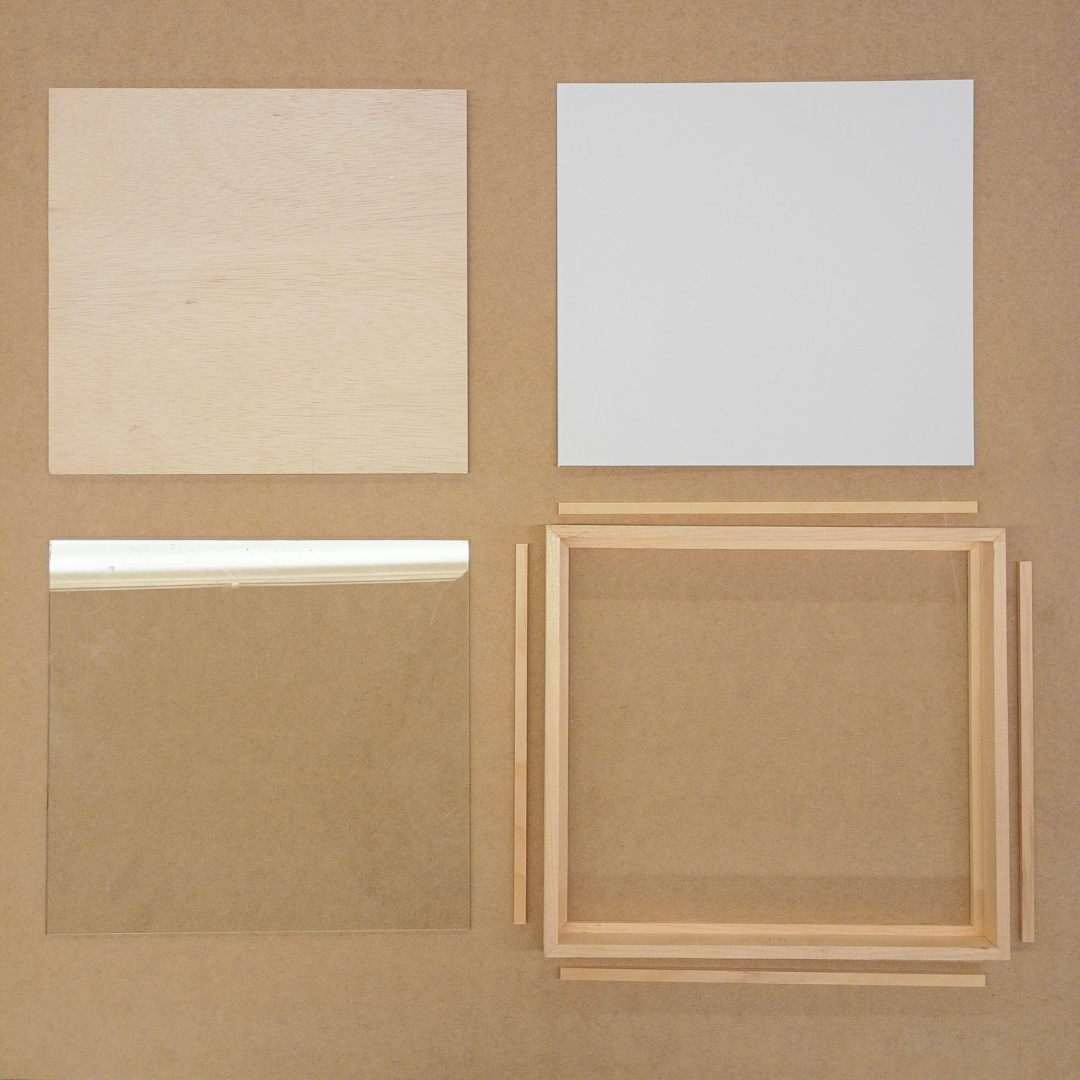 An overhead view displays the disassembled components of a wooden shadow box frame, including a solid wood backing, a white mat board, a pane of glass, and the rectangular frame structure with its interior spacers.