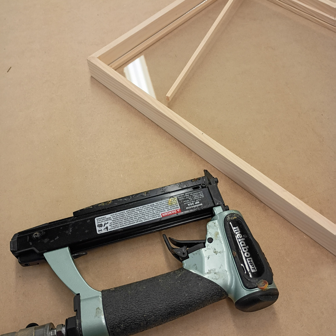 A Metabo HPT pneumatic pin nailer rests on a workbench next to a partially assembled wooden picture frame.