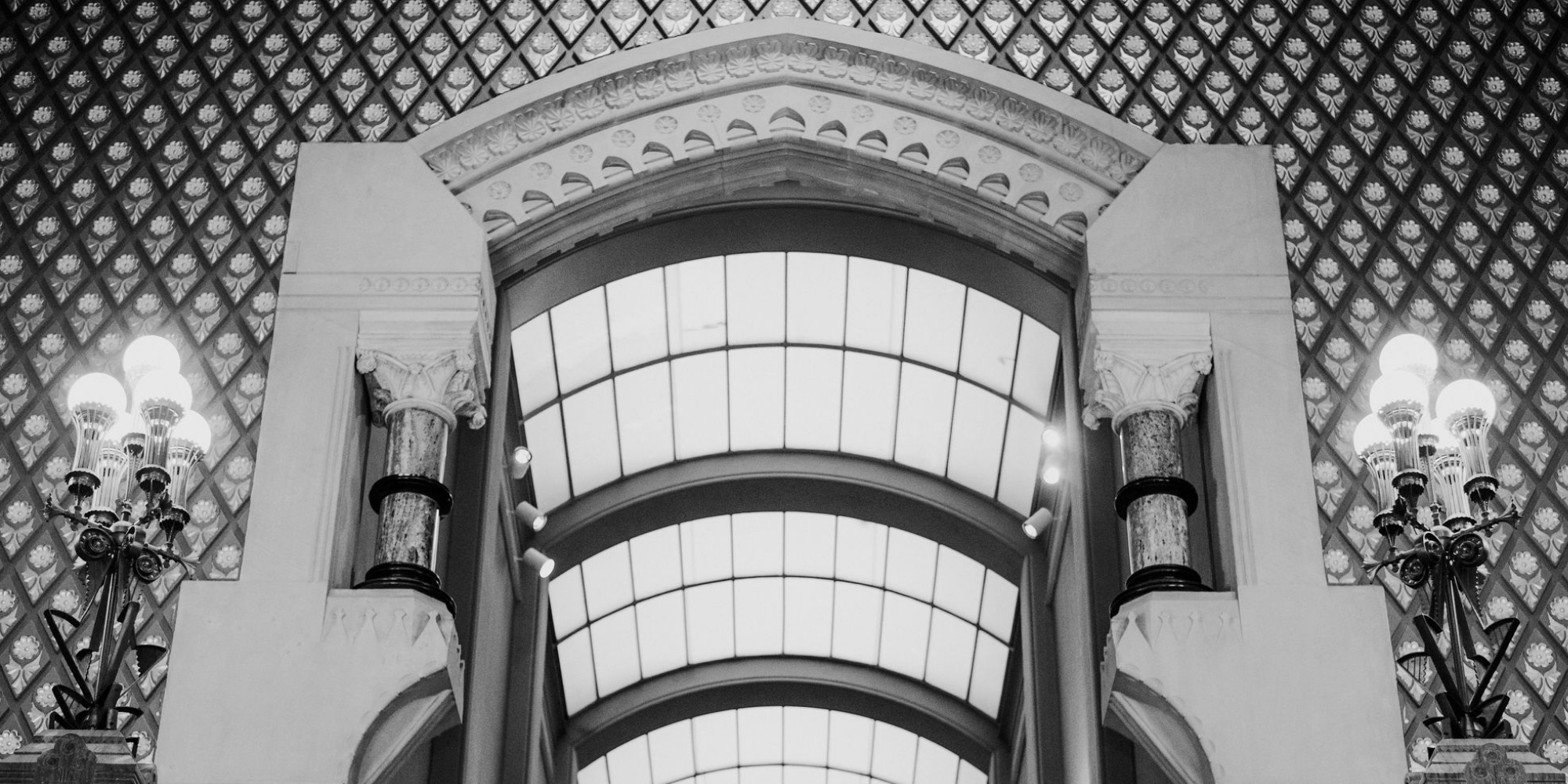 a black and white photograph of interior space of the Historic Landmark Building, featuring a large, ornate arched window flanked by decorative, multi-bulb lamp posts.