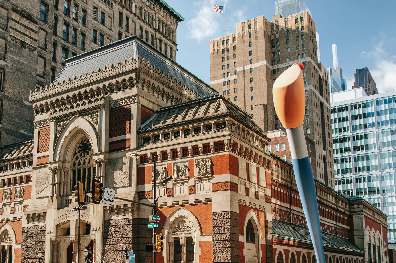 Historic building with ornate architecture featuring red and beige bricks, arched windows, and sculpted details. A giant paintbrush sculpture stands prominently in the foreground, symbolizing art and creativity against a backdrop of modern skyscrapers.