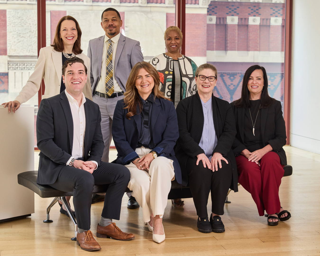 A professional group portrait of six colleagues in business attire posing together indoors, with three seated on a bench and three standing behind them.