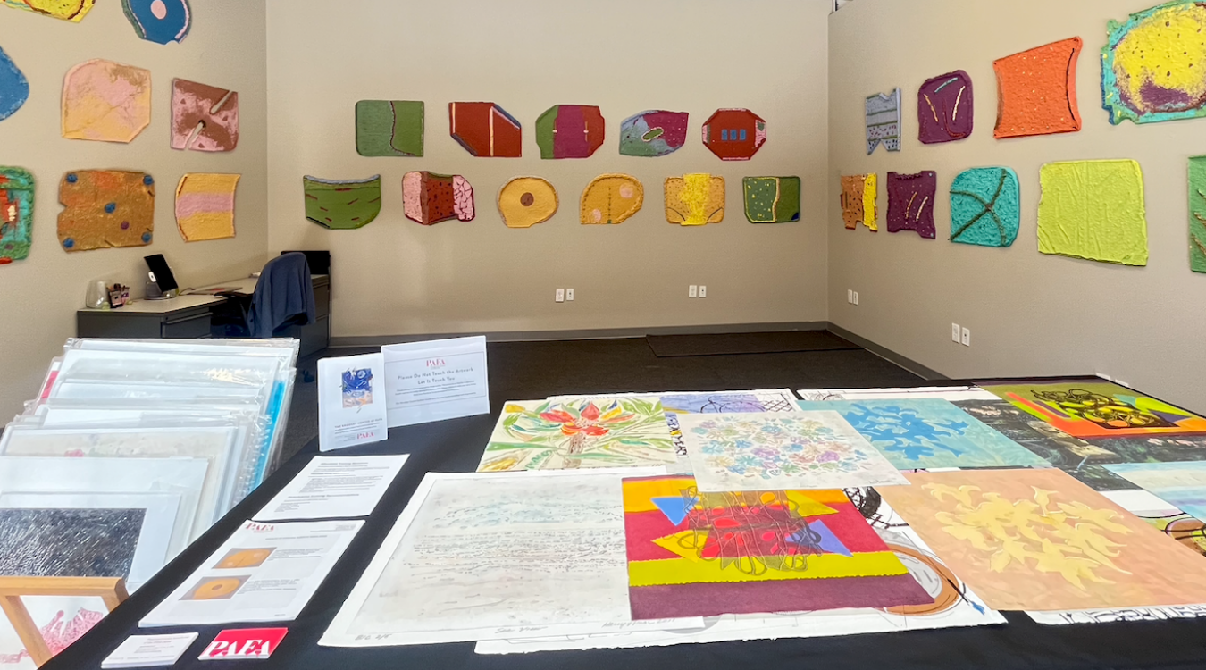 This image appears to show an art gallery or community exhibition space featuring framed textile or fiber art wall hangings, works on paper, and assorted mixed-media collage pieces laid out on display tables.