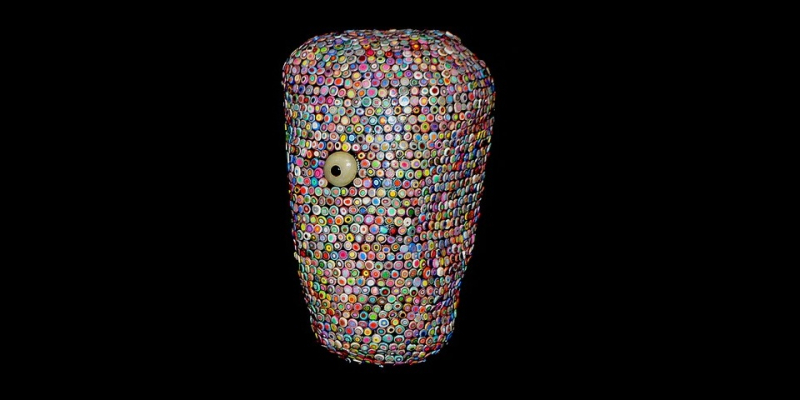  Mixed-media bust intricately encrusted with dense layers of multi-colored bottle caps and buttons, featuring a prominent, singular off-center eye.