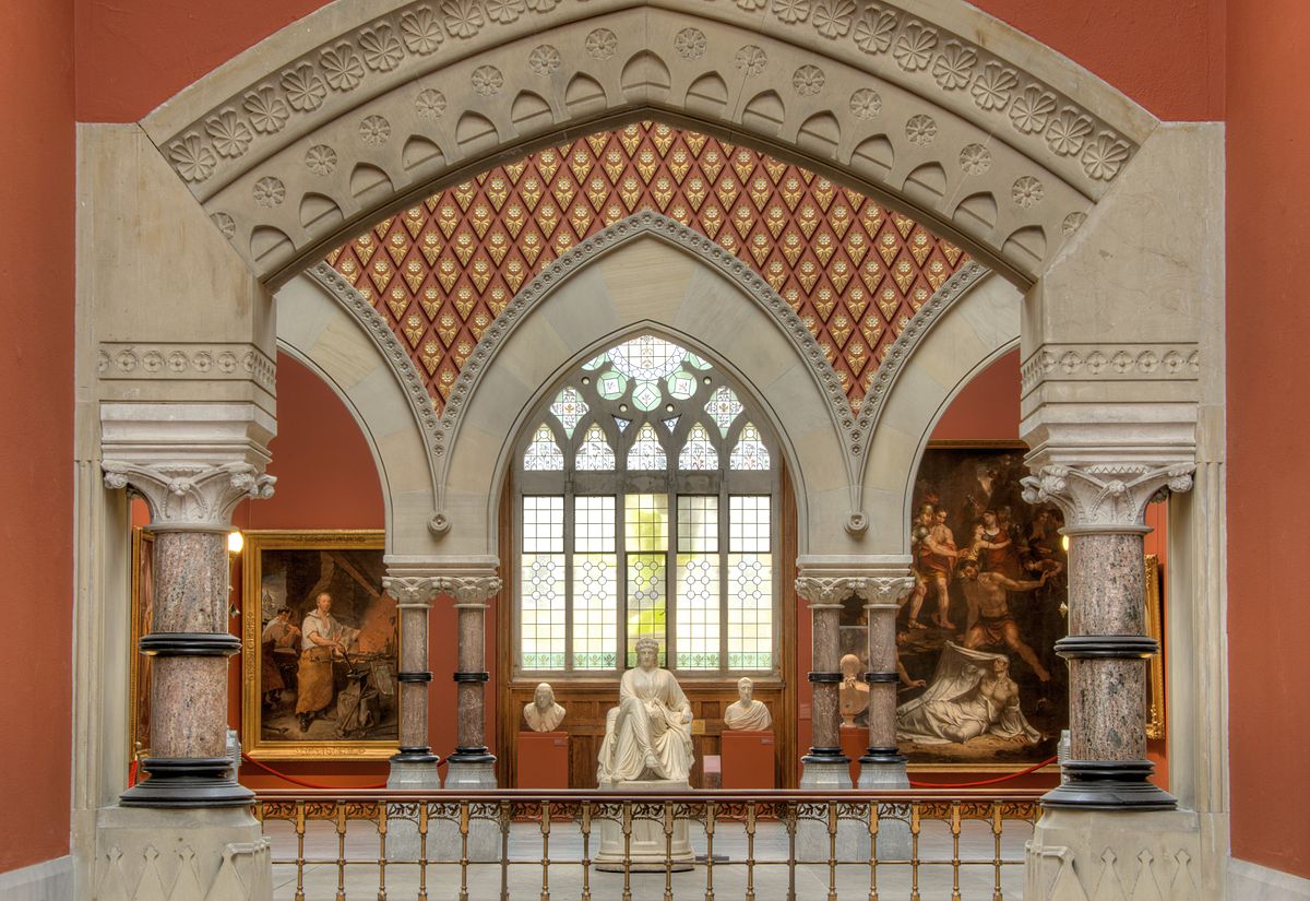 Gothic arched gallery with ornate columns, red and gold ceiling; three marble statues in front of a stained glass window, flanked by large paintings.