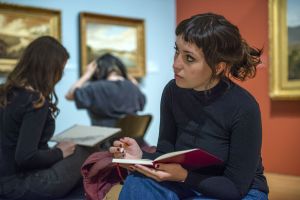 Photo of three women sitting in a gallery. The woman closest to the camera has dark hair with a short pony-tail and is wearing a black top. She is holding an open notebook and a pencil, looking up and away from the camera. 