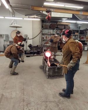 Photo of four people dressed in heavy brown protective clothing, goggles and gloves, handling the pour of molten bronze into openings in a dark gray solid casing. The cauldron with the bronze is controlled by a pully above.