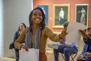 photo of Misty Sol smiling and handing a piece of paper to someone not seen in the photo. There are three people sitting on stools visible in the background. There are large painted portraits on red walls in the far back. 