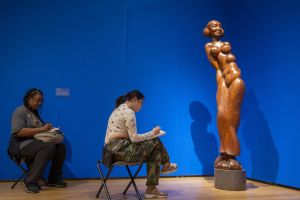 photo of two women sitting in art gallery sketching on small pads of paper. A tall wood figurative sculpture is on the right side of the frame.