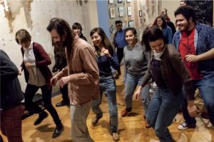 Photo of at least 14 people, most appear to be in their 20s,  wearing casual clothes dancing in a room with wooden floors.
