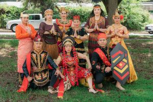 Group photo of the Modero company. There are nine dancers, three kneeling and six standing, holding a dance pose. All the dancers are wearing intricate and colorful costumes. The photo was taken outdoors, with trees and cars in the background.  