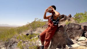 image of a person sitting a top of a rock on a hill holding a wood and metal kilimba (thumb piano) over their face. 