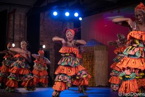 Photo of five dancers in bright orange African print costumes. The troupe is on a stage with three blue stage lights abover their heads. 