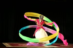 A single dancer in a white embroidered dress and red embroidered jacket, with detailed face painting and headress, dancing on a stage, twirling long colorful ribbons, one in each hand, swirling around her body. 