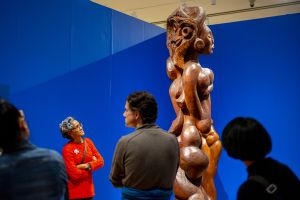 Photo of a a team of five people handling heavy equipment to pour molten bronze into molds. Behind the team stand five people looking intently at the pour in front of them. 
