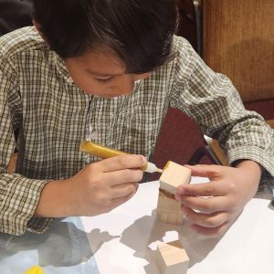 This image shows a child crafting with small wooden blocks and gold-colored glue.