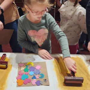 A child engaging in an art activity that involves sand and a roller. The person is wearing a long-sleeved shirt with the word "BROOKLYN" written across a heart graphic.